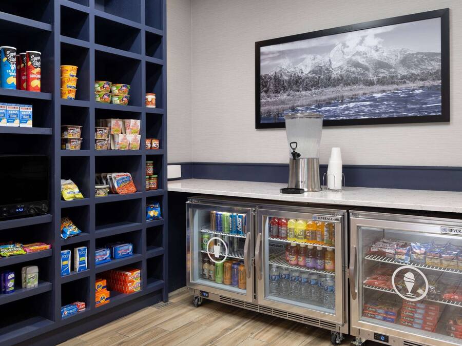 A kitchenette with shelves stocked with snacks and beverages, a refrigerator with drinks, and a framed picture of a snowy landscape on the wall.