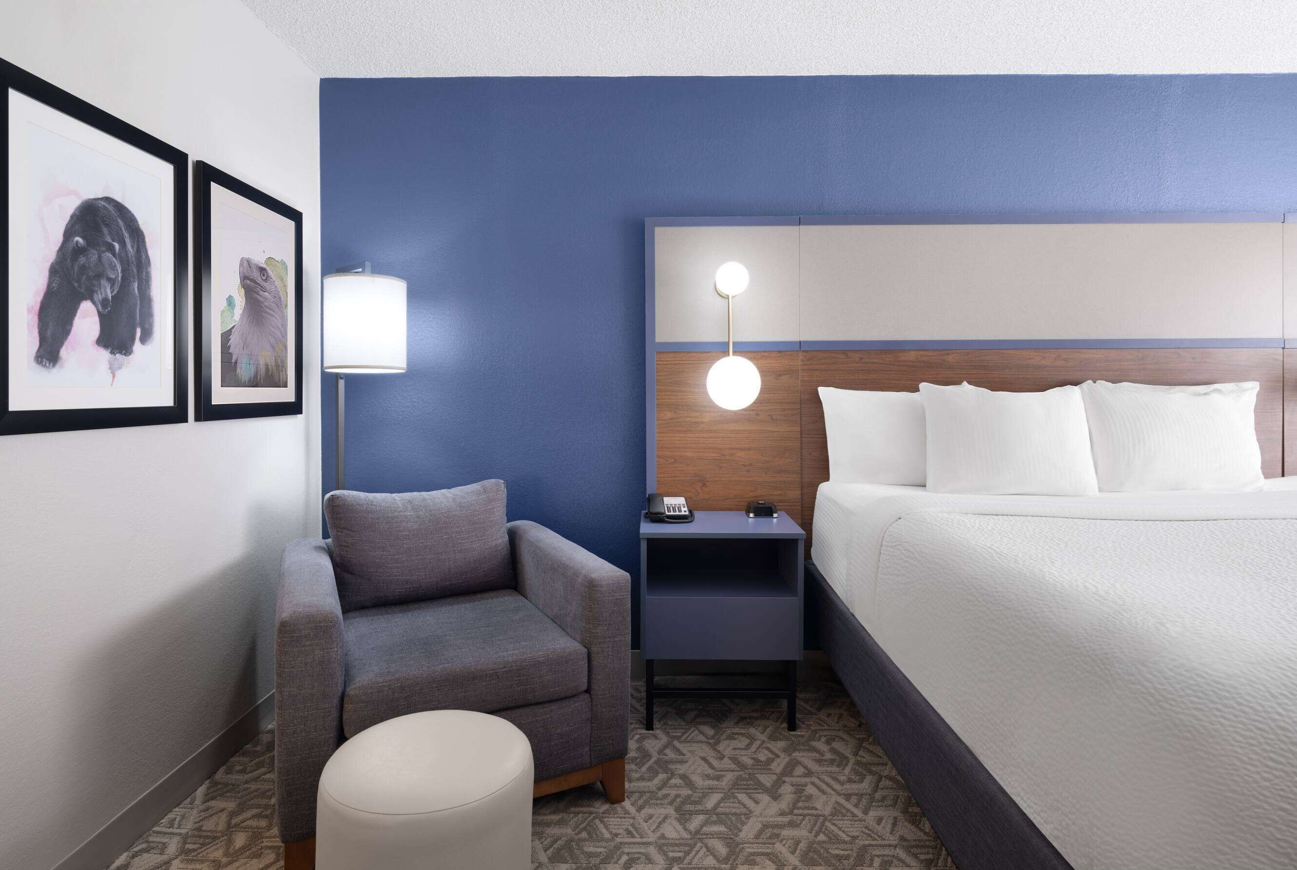 A hotel room with a bed, a gray armchair, a small table, a lamp, and two framed pictures on the wall.