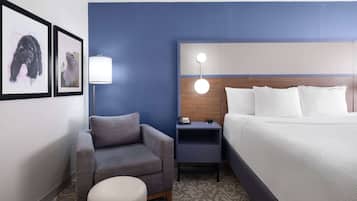A hotel room with a bed, a gray armchair, a small table, a lamp, and two framed pictures on the wall.