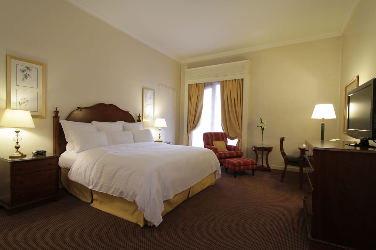 Habitación de hotel con una cama grande, mesitas de noche con lámparas, un escritorio con televisor y una silla.