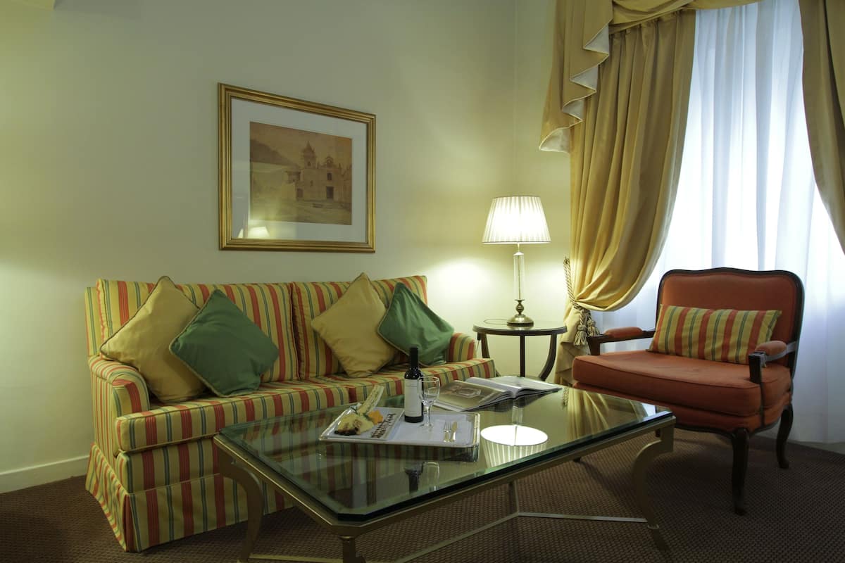 Habitación de hotel con un sofá a rayas, una mesa de centro de vidrio, una silla, una lámpara y un cuadro en la pared.