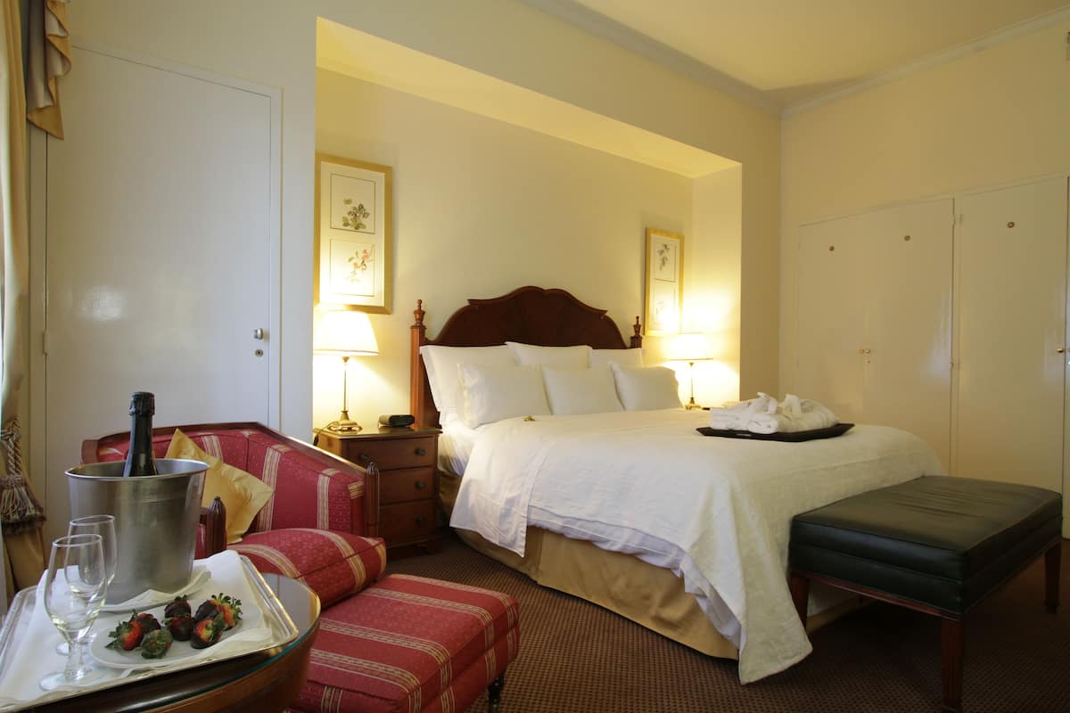 Una habitación de hotel con una cama, mesitas de noche, un sillón rojo, una pequeña mesa con una bandeja de fresas y una botella de champán, y un banco.