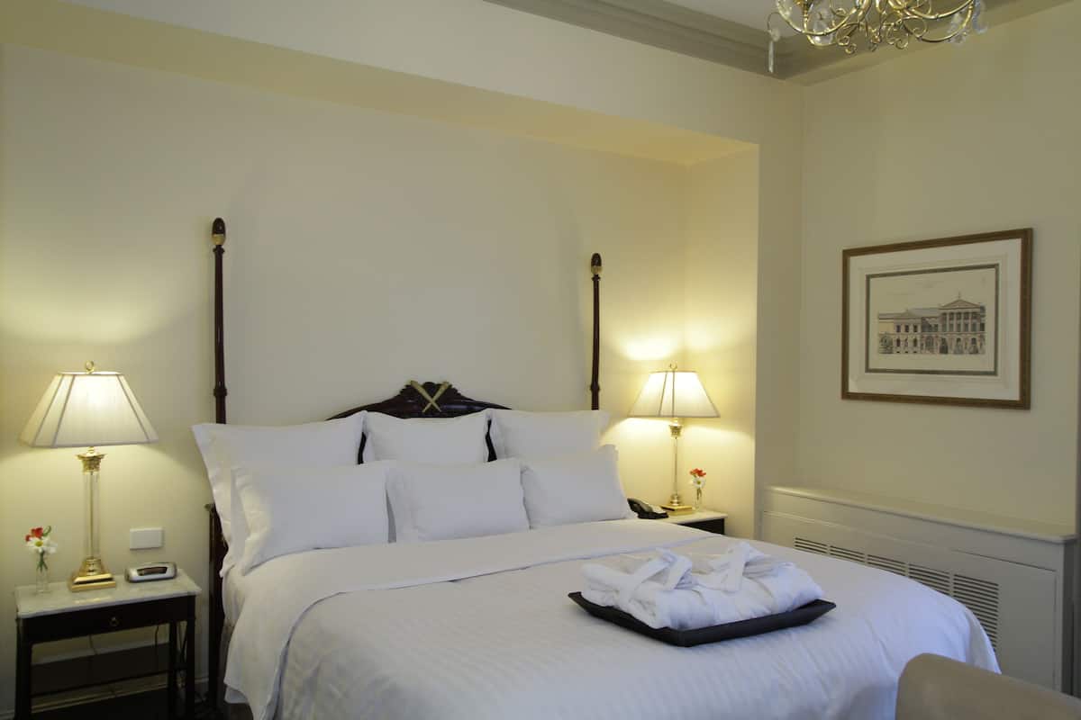 Una cama bien hecha con sábanas blancas, dos lámparas de noche, un cuadro en la pared y una mesita con un jarrón de flores.