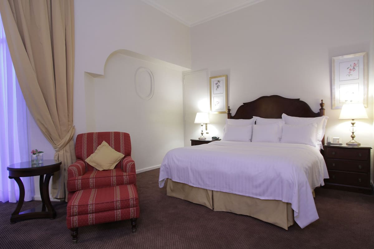 Una habitación de hotel con una cama, un sillón rojo a cuadros, una mesita de noche con una lámpara y una pequeña mesa con un florero.