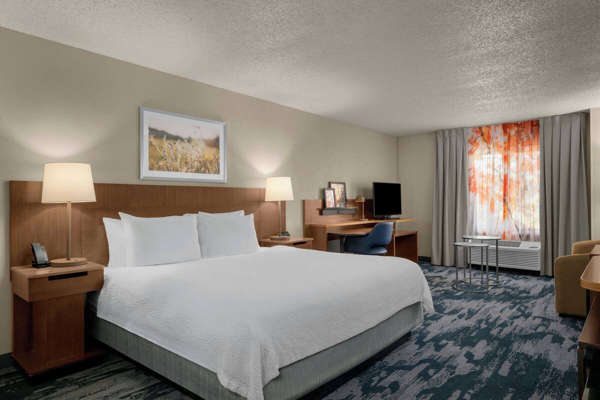 A hotel room with a large bed, two bedside tables with lamps, a desk with a chair, a television, and a window with curtains.