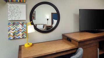 A hotel room with a wooden desk, a television, a mirror, a lamp, and artwork on the wall.