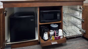 A compact kitchenette with a black refrigerator, a microwave, a coffee maker, and a small shelf with various items.