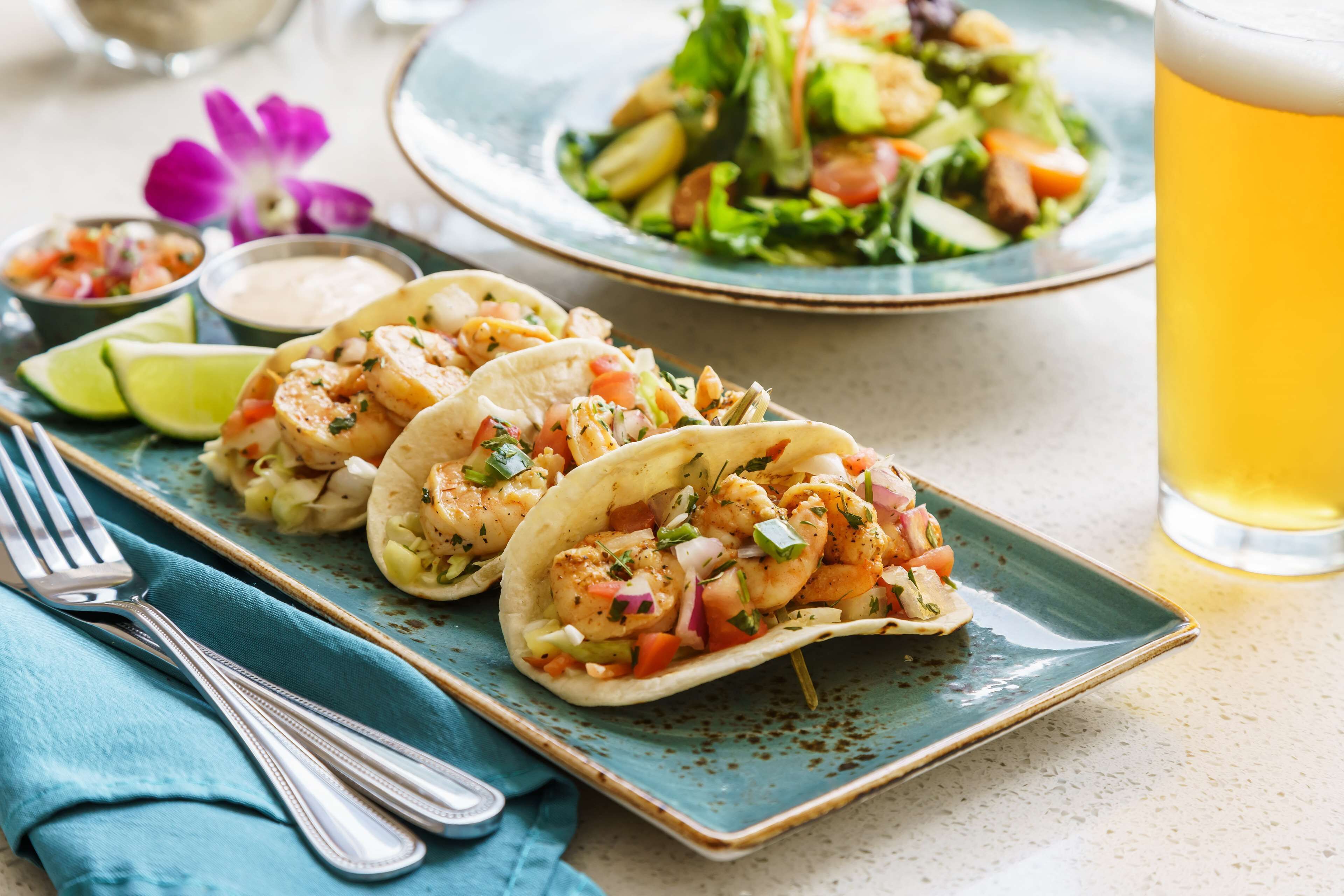 Three shrimp tacos on a plate, a glass of beer, and a side salad.