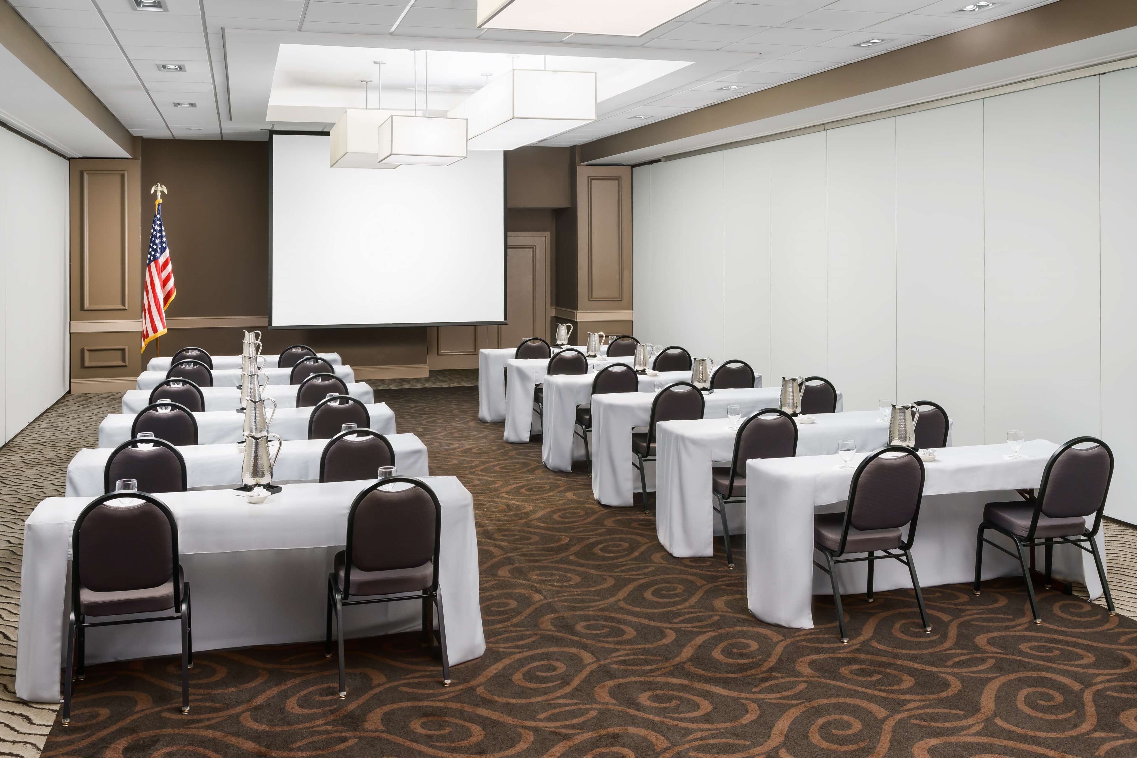 A conference room with multiple tables and chairs, a projector screen, and an American flag.