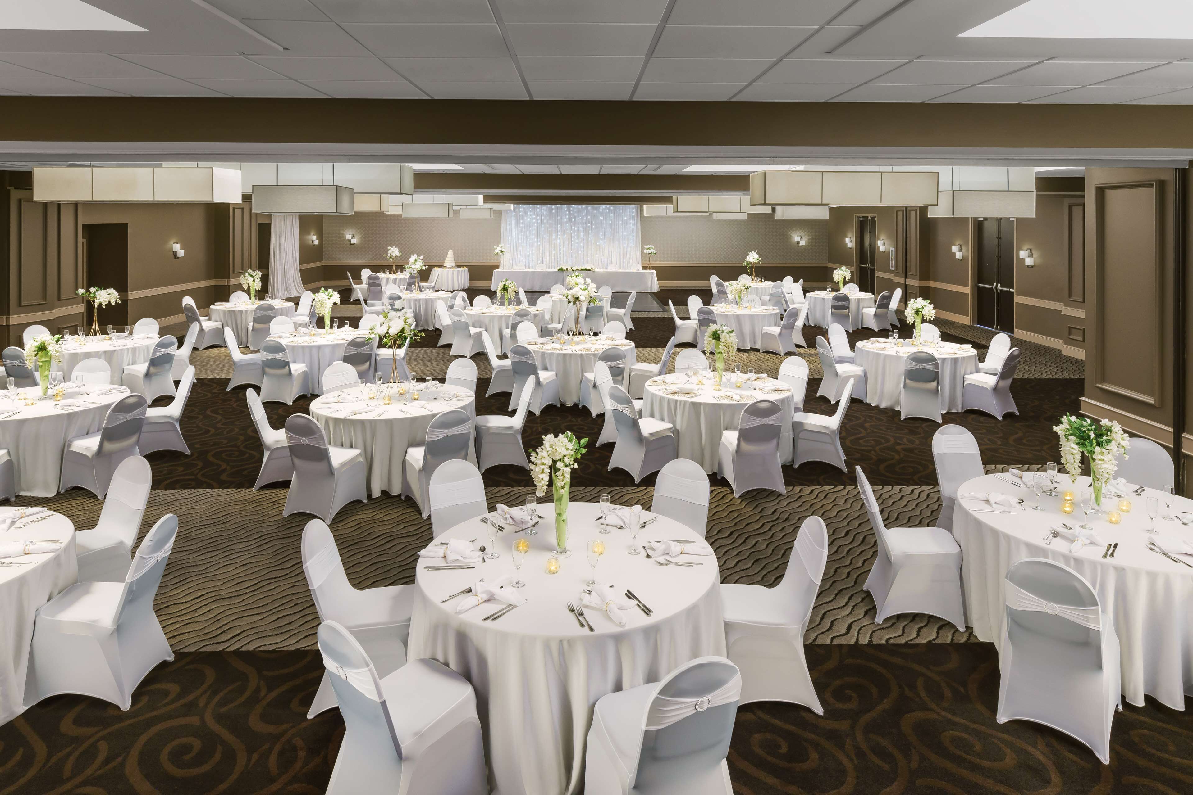 A banquet hall with multiple round tables, each set with white tablecloths, chairs, and floral centerpieces.