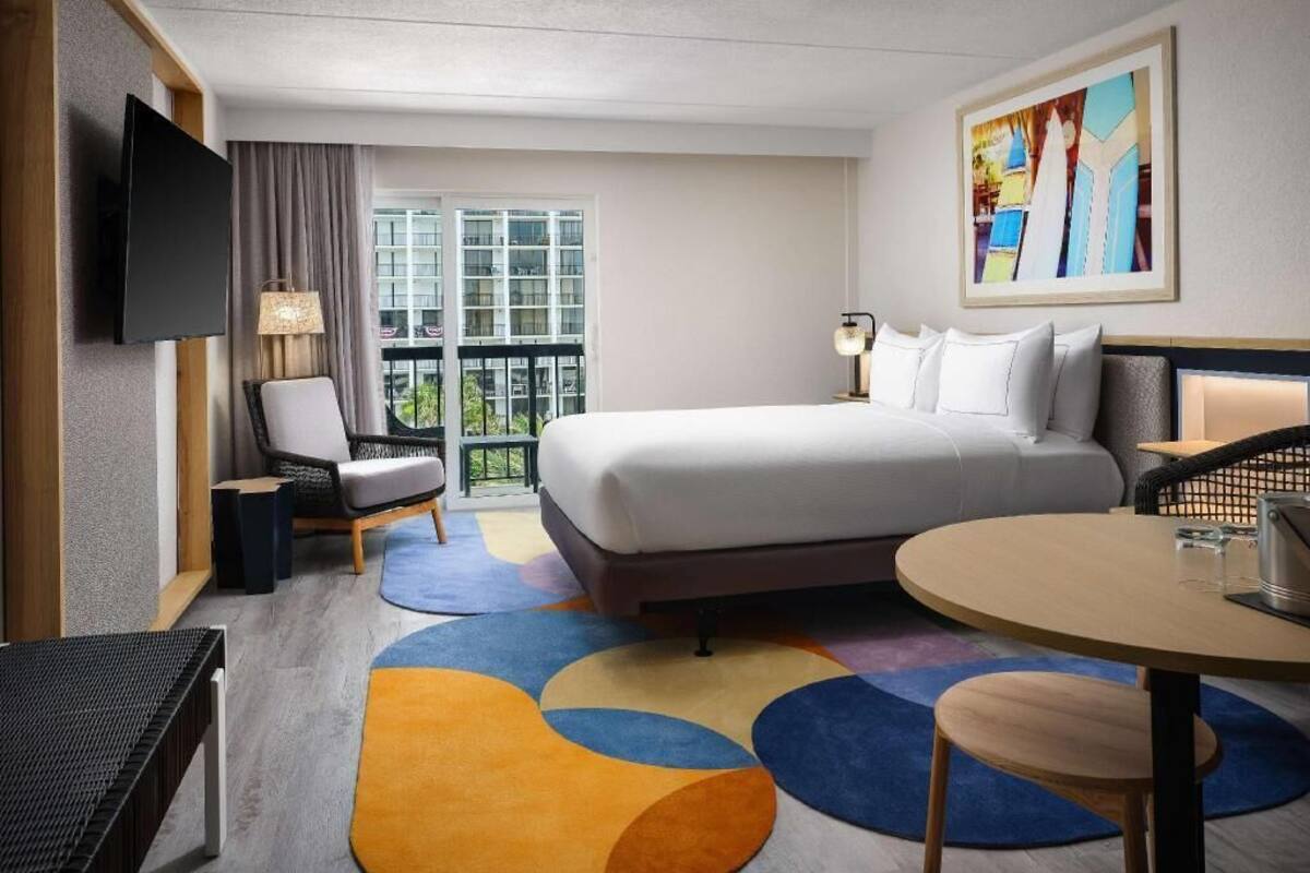 Habitación de hotel con cama, escritorio, silla, televisor, ventana con vistas y un cuadro en la pared.