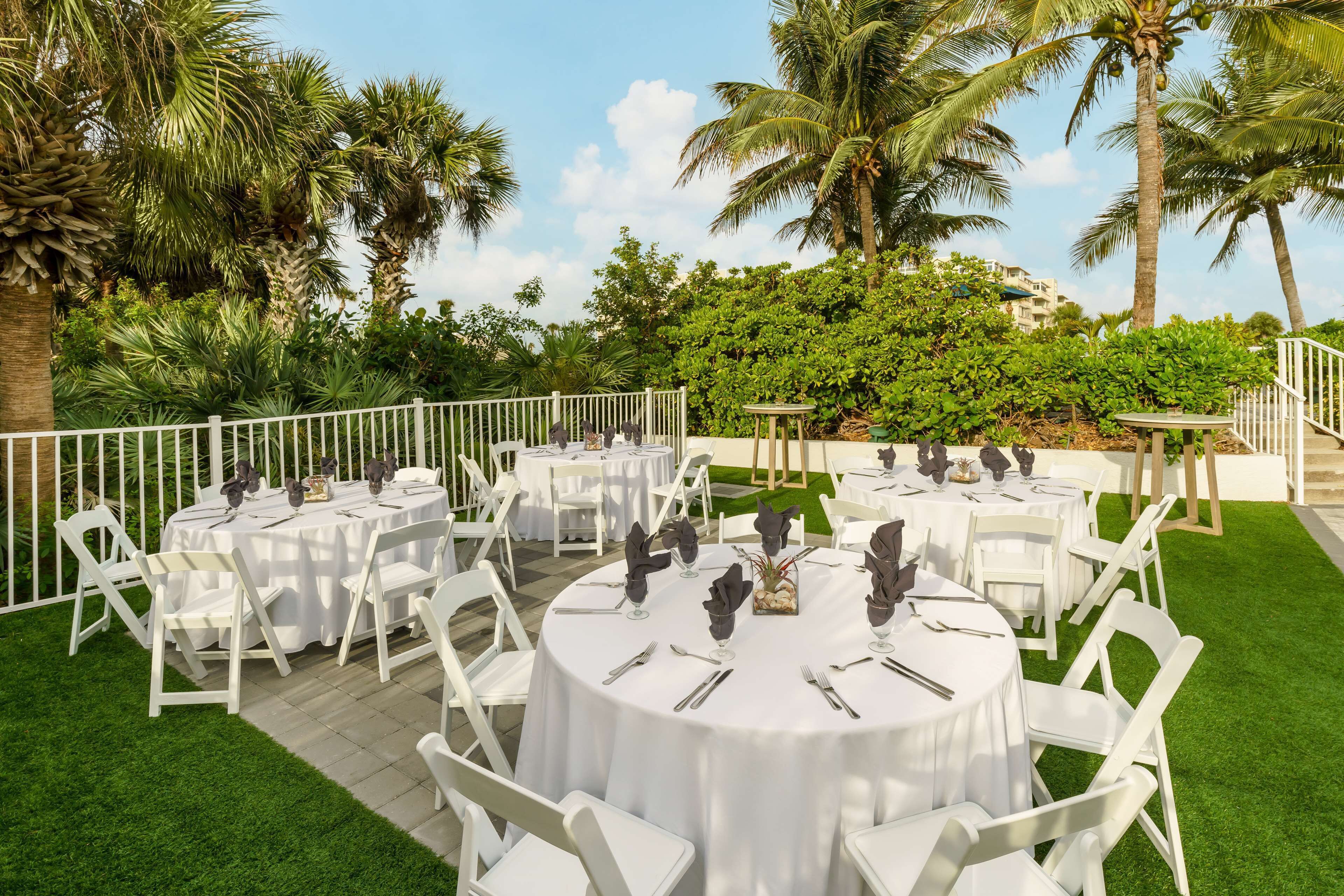 An outdoor dining area with round tables and white chairs, surrounded by palm trees and greenery.