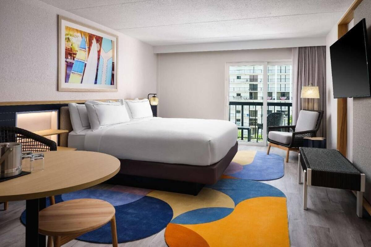 Habitación de hotel con cama, escritorio, silla, televisor y ventana con vista a edificios.
