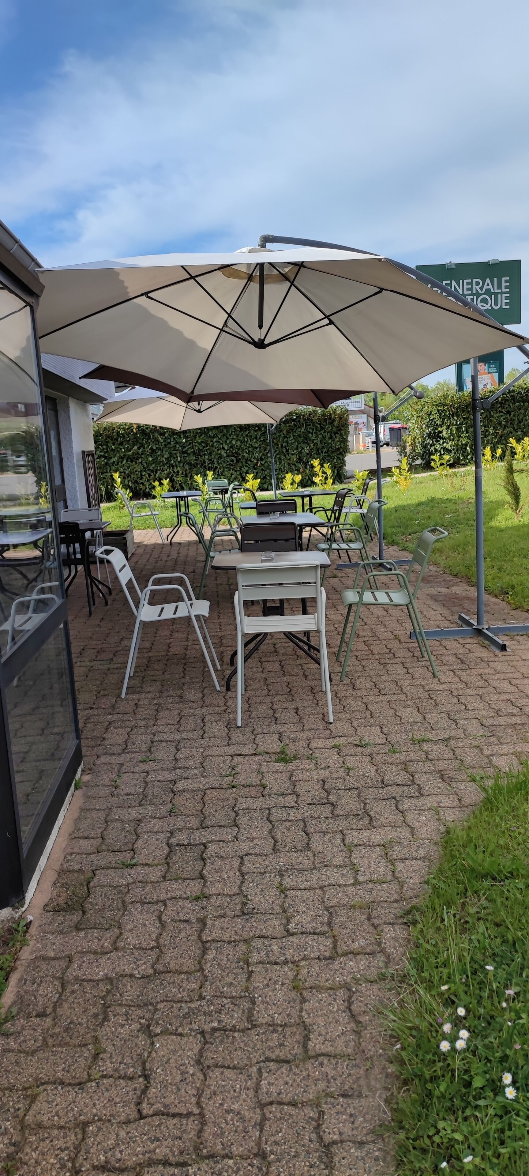 An outdoor patio with tables and chairs under a large umbrella, surrounded by greenery and a paved walkway.