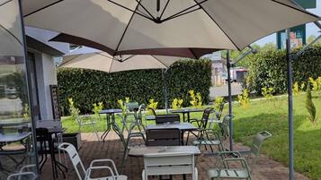 An outdoor patio with tables and chairs under a large umbrella, surrounded by greenery and a paved walkway.