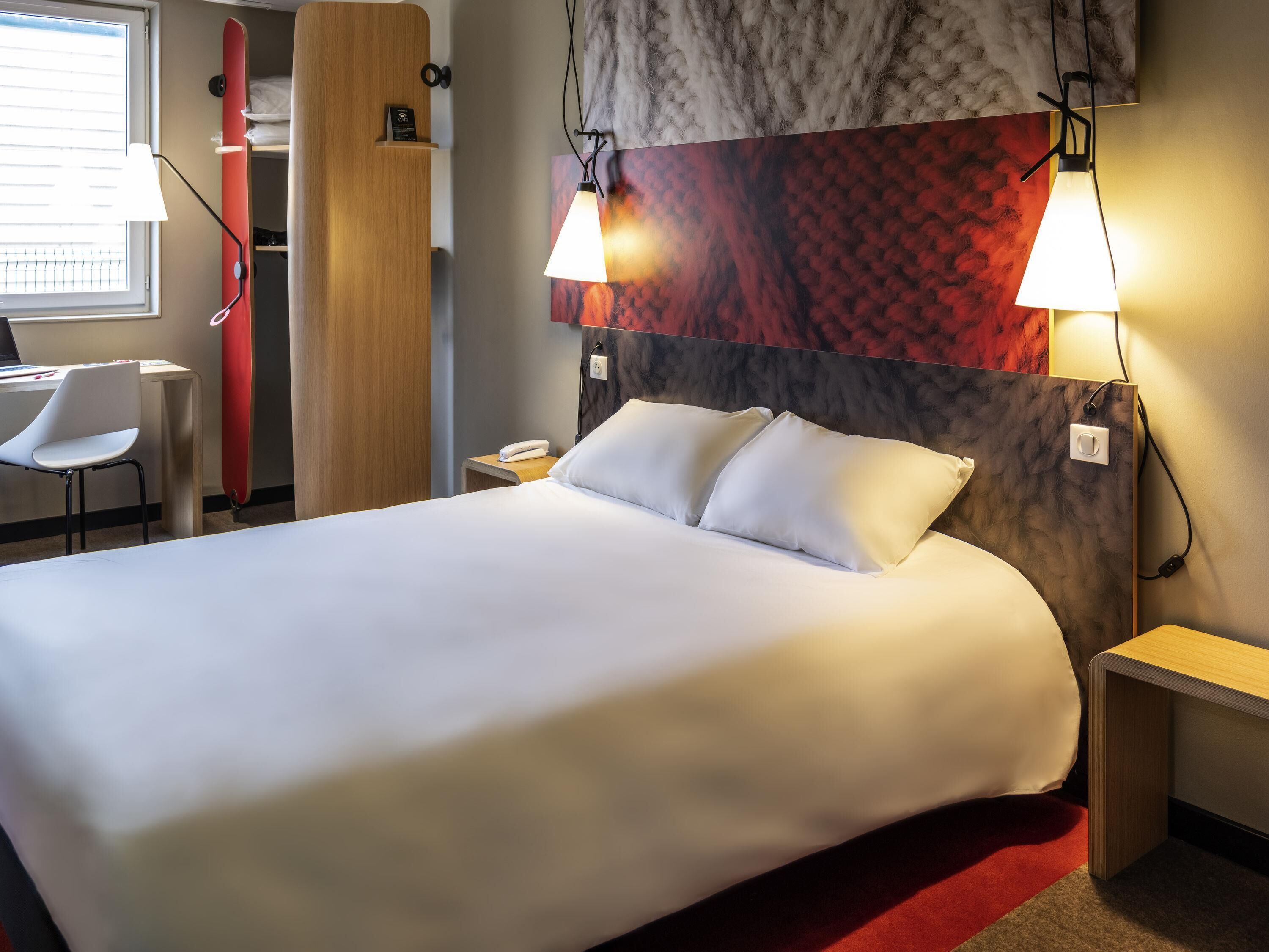 Habitación de hotel con una cama grande, mesitas de noche, un escritorio con silla, un armario y una ventana con persianas.
