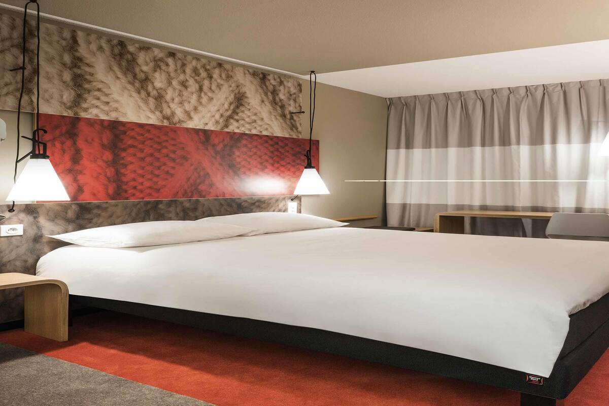 Quarto de hotel com cama grande, duas luminárias de cabeceira, mesa de cabeceira de madeira, uma pequena mesa e uma janela com cortinas.