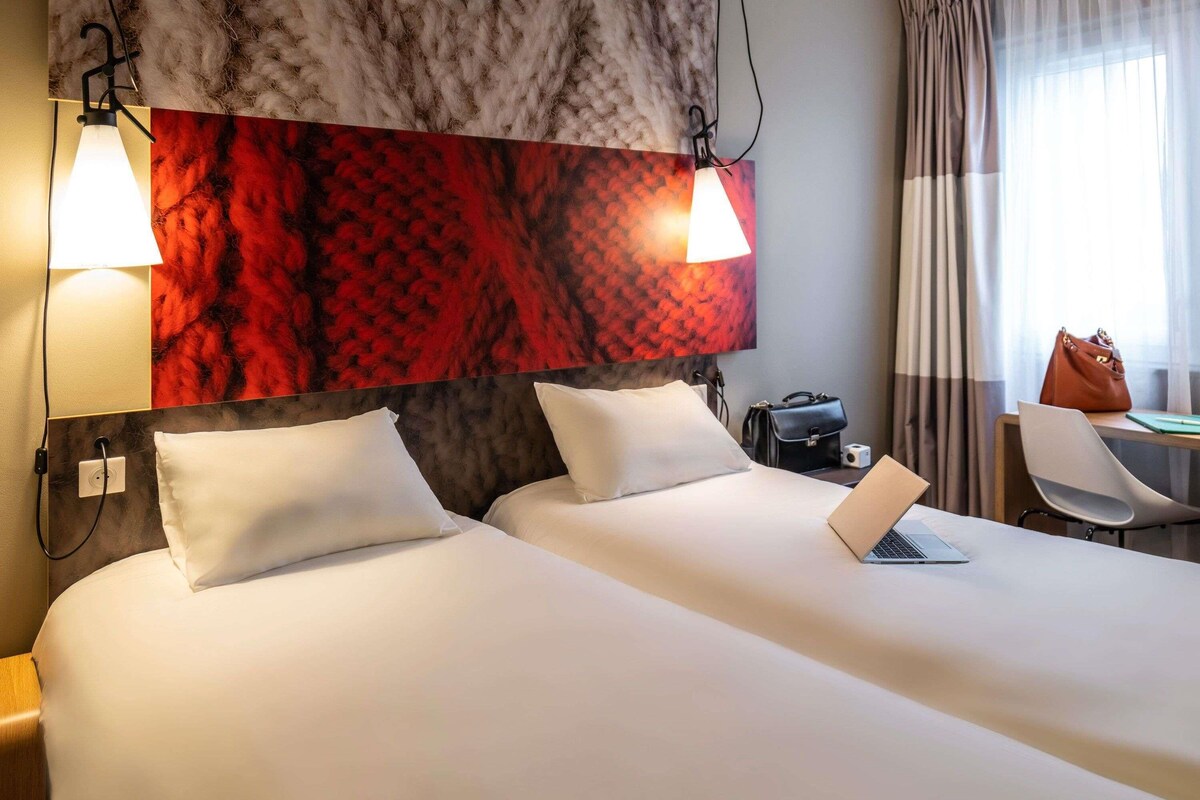 A hotel room with a large bed, two bedside lamps, a desk with a laptop, a chair, and a handbag.