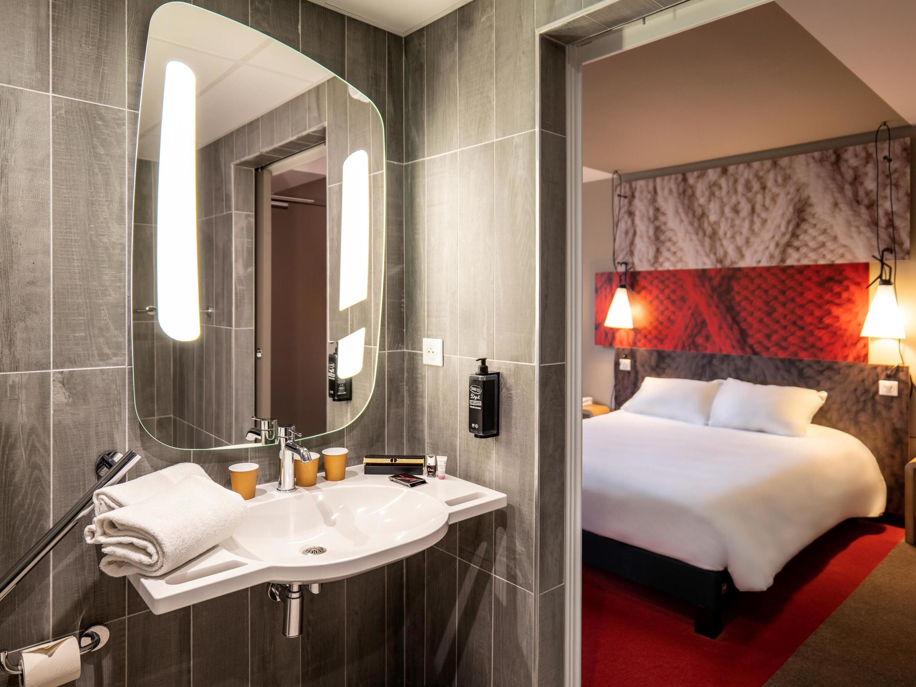 A hotel room with a modern bathroom, a large mirror, a sink, and a bed with a red accent wall.