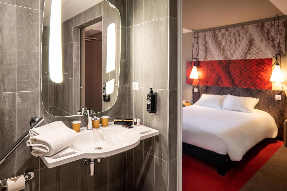 A hotel room with a modern bathroom, a large mirror, a sink, and a bed with a red accent wall.