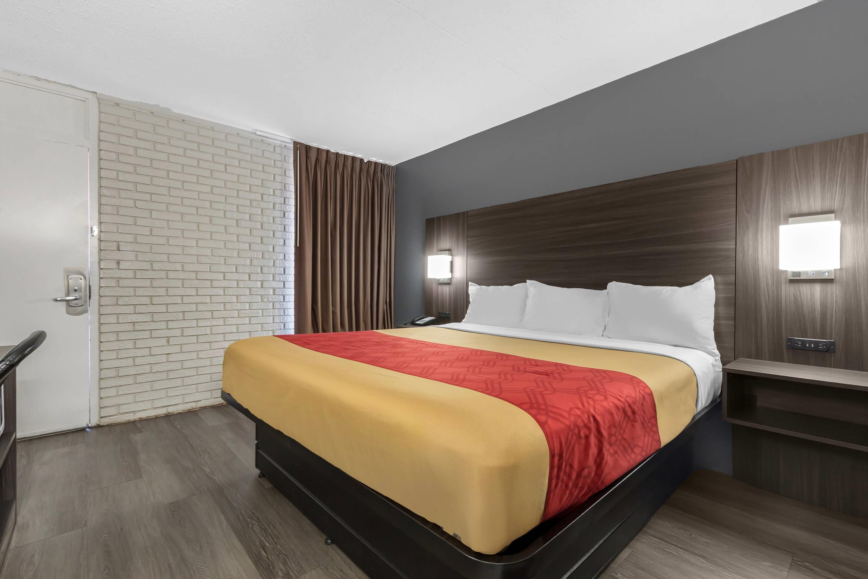 A hotel room with a bed, a door, a brick wall, a window with curtains, and a nightstand with a lamp.
