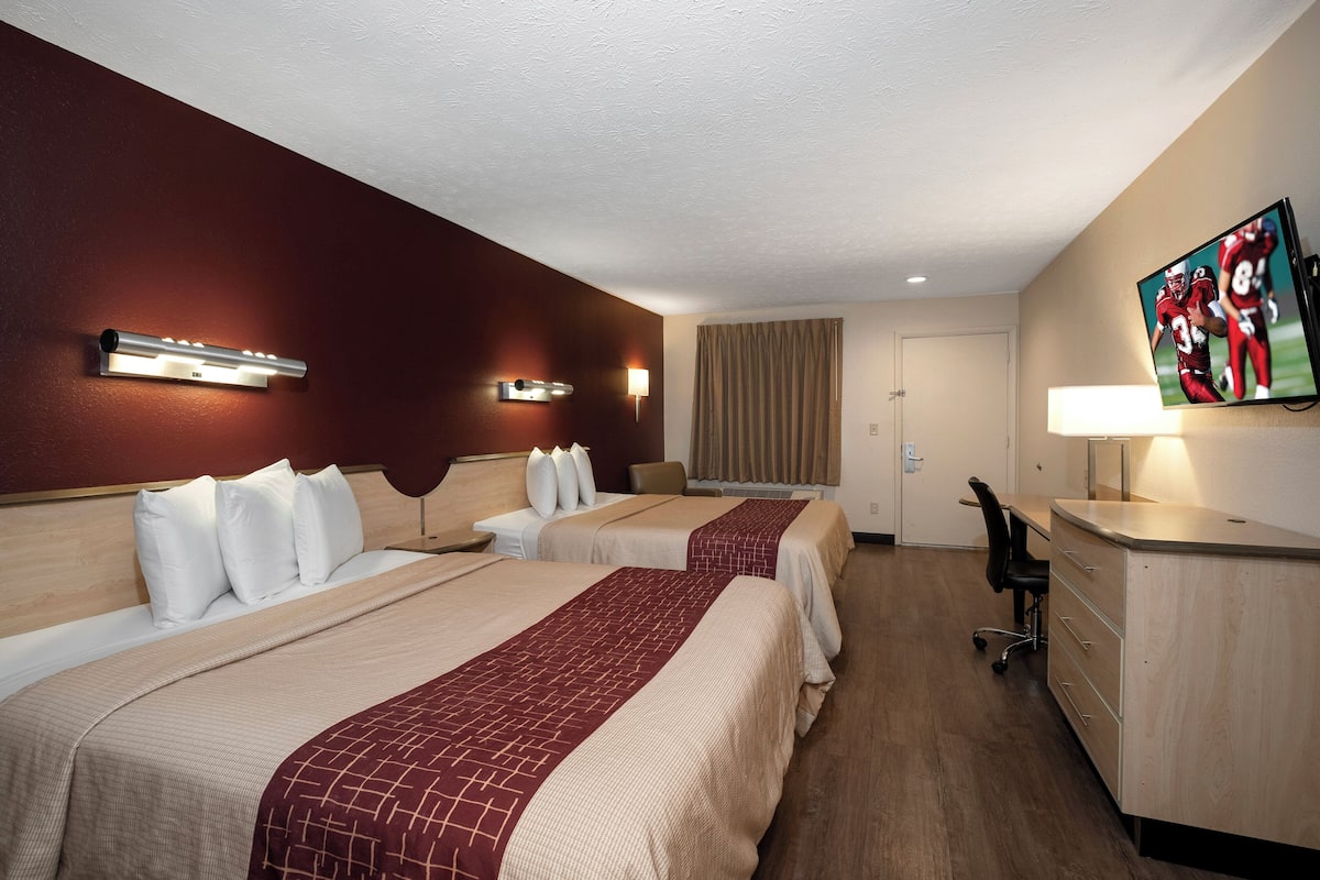 A hotel room with two beds, a desk, a chair, a TV, and a lamp.