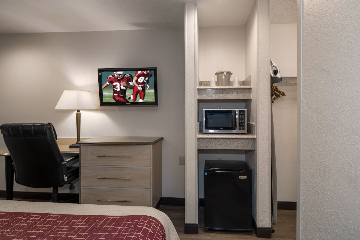 A hotel room with a bed, a desk with a chair, a television displaying a sports game, a microwave, and a refrigerator.