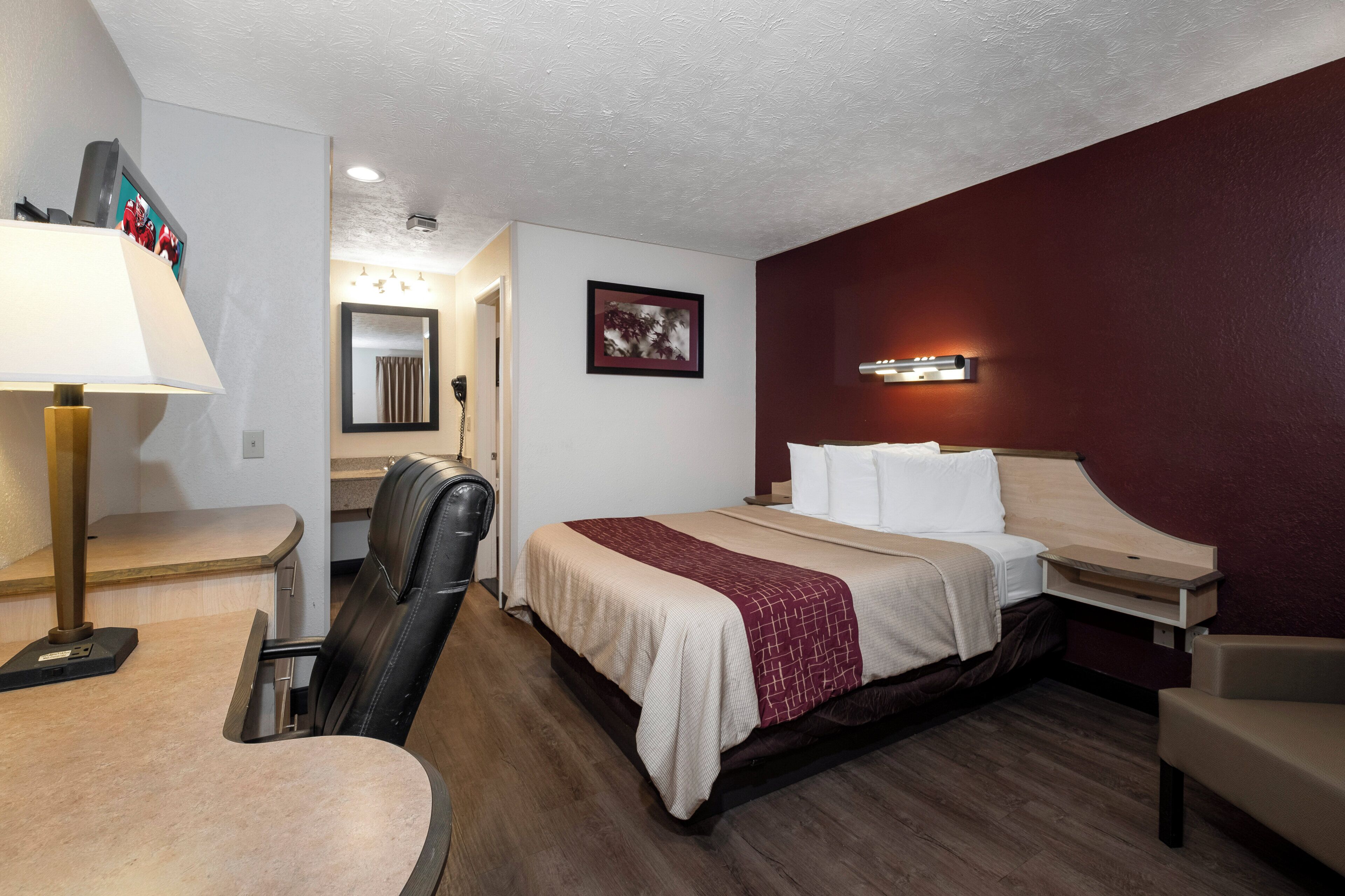 A hotel room with a bed, a desk, a chair, a lamp, a mirror, and a picture on the wall.