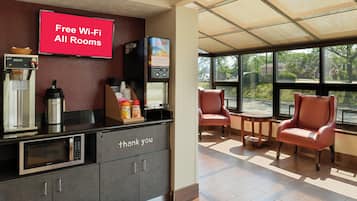 A hotel reception area with a 'Free Wi-Fi All Rooms' sign, a juice machine, a coffee machine, and a 'thank you' sign on the counter.