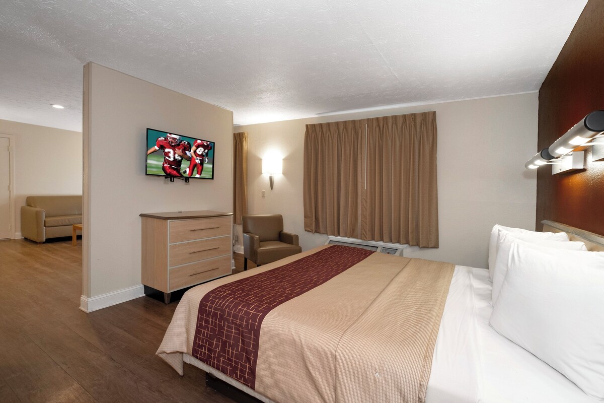 A hotel room with a bed, a TV mounted on the wall, a desk, a chair, and a window with curtains.