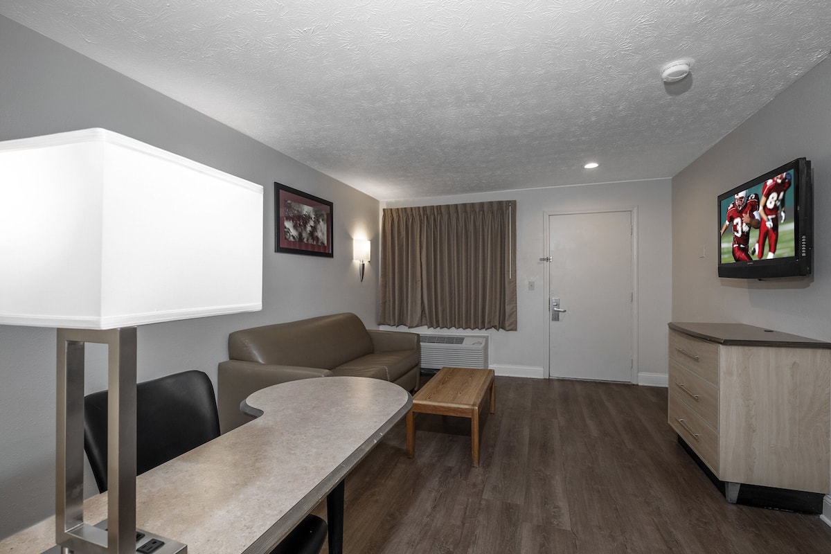 A modern hotel room with a dining table, sofa, TV, and a small wooden cabinet.