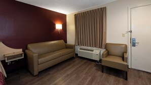 A hotel room with a sofa, a chair, a wall-mounted heater, a curtain, and a door. - Red Roof Inn Indianapolis - Greenwood (Greenwood)