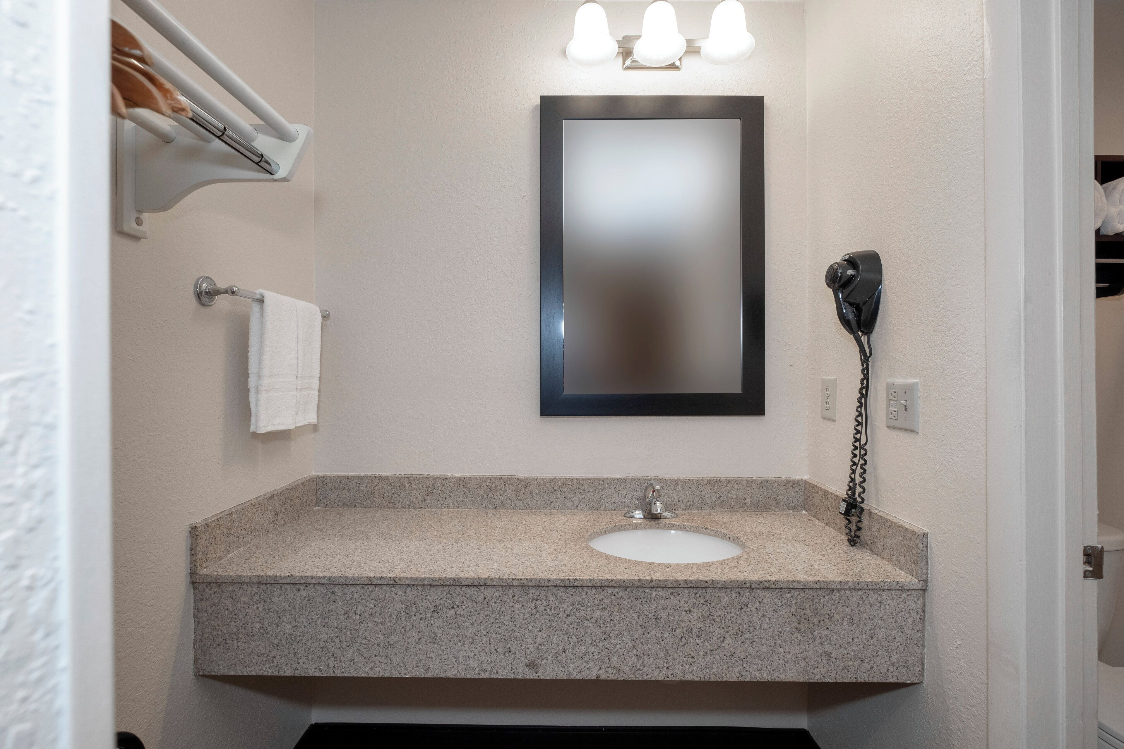 Une salle de bain avec un lavabo doté d’un plan de travail en granit, un miroir, un porte-serviettes et un sèche-cheveux fixé au mur.