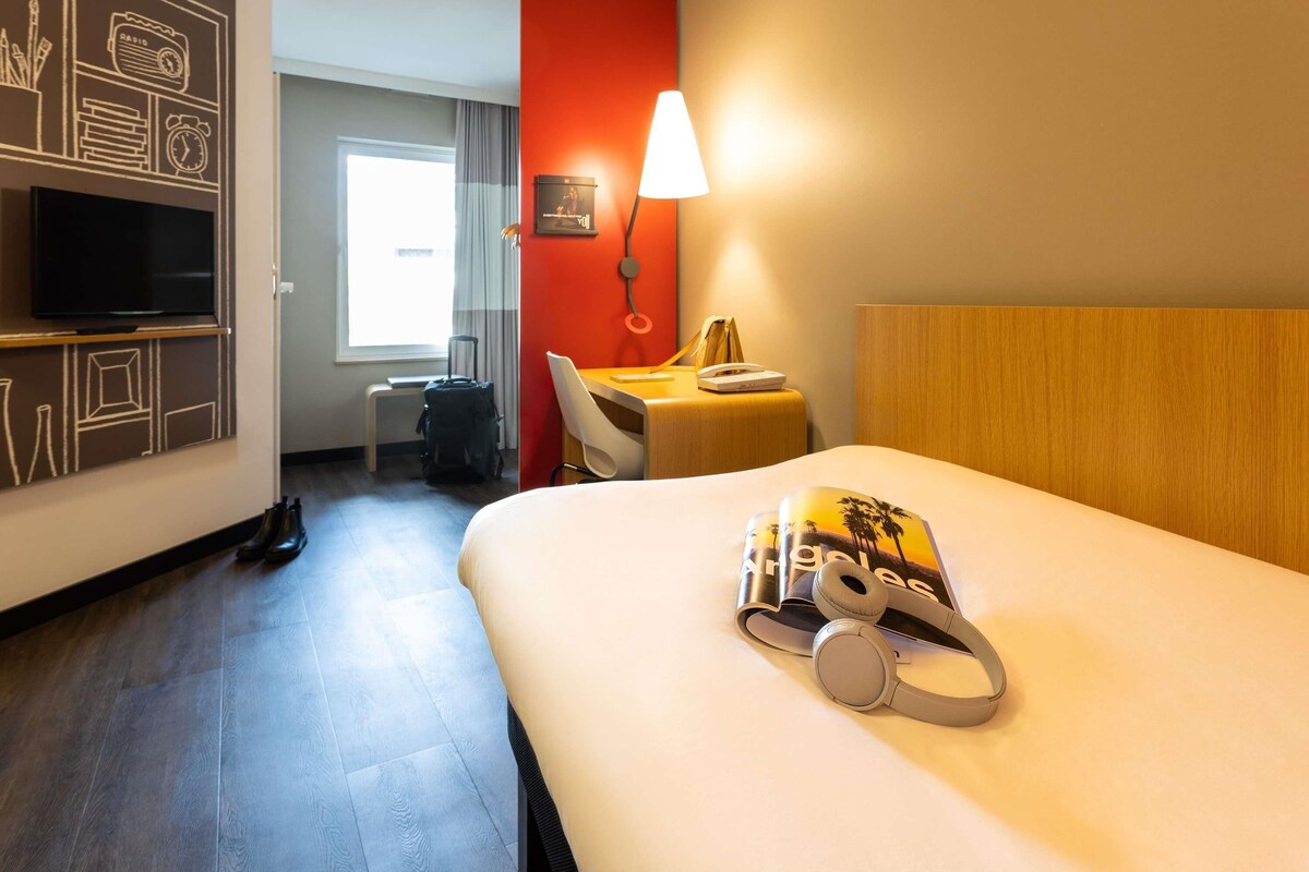 A hotel room with a bed, a desk, a chair, a TV, a lamp, and a suitcase.