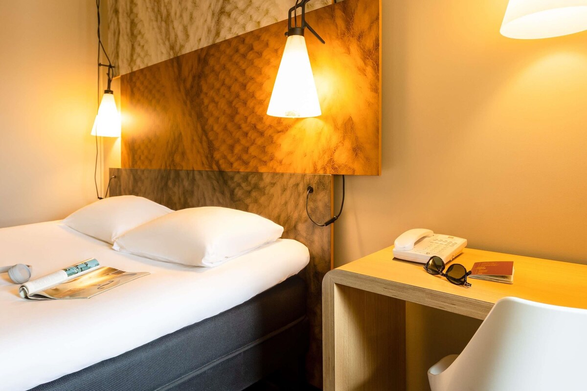 A hotel room with a bed, a bedside table with a phone and glasses, and a magazine on the bed.