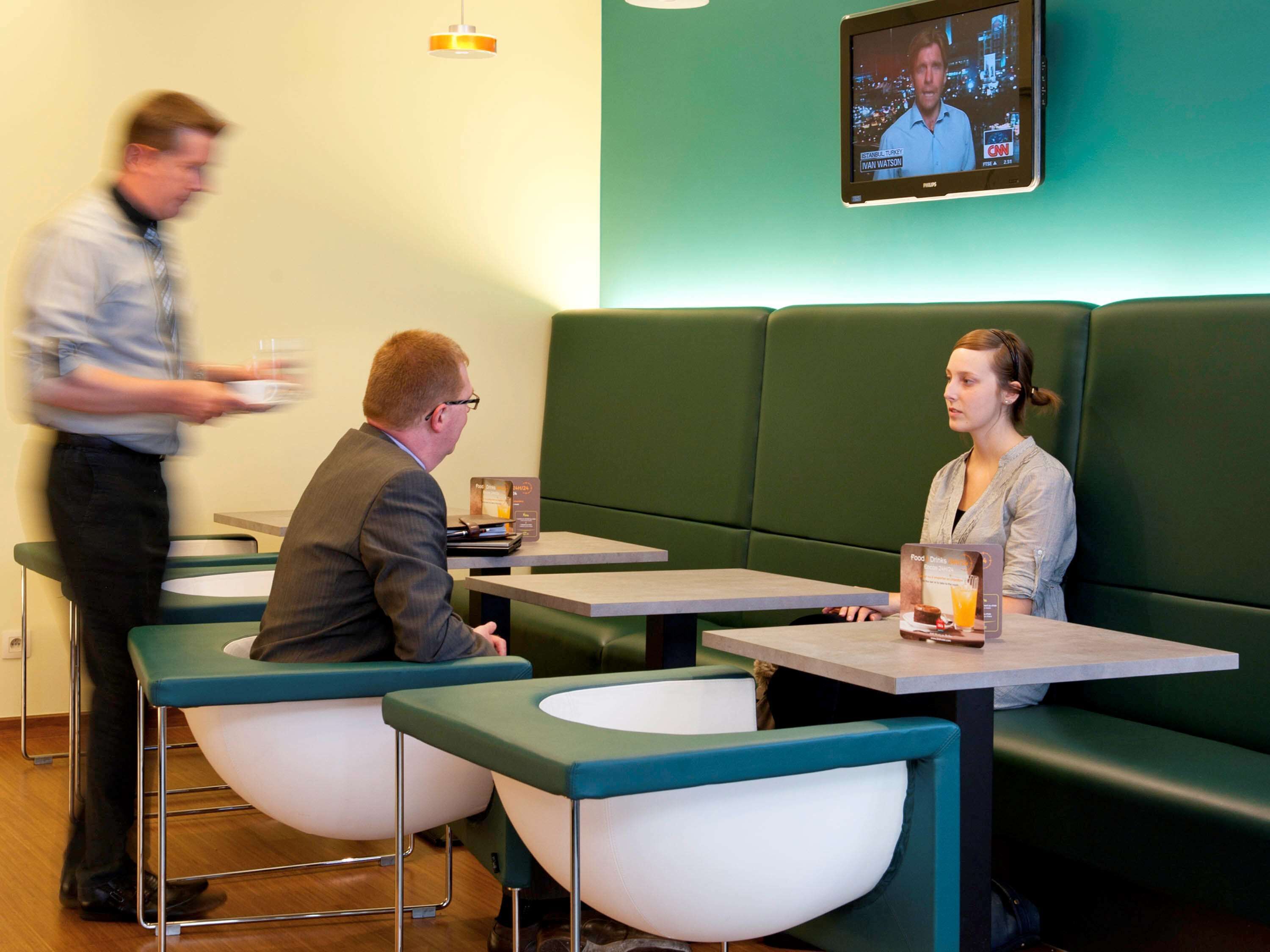 Een modern café met groene zitplaatsen en tafels, een televisiescherm waarop een nieuwsuitzending draait, en een menukaart die op tafel staat.