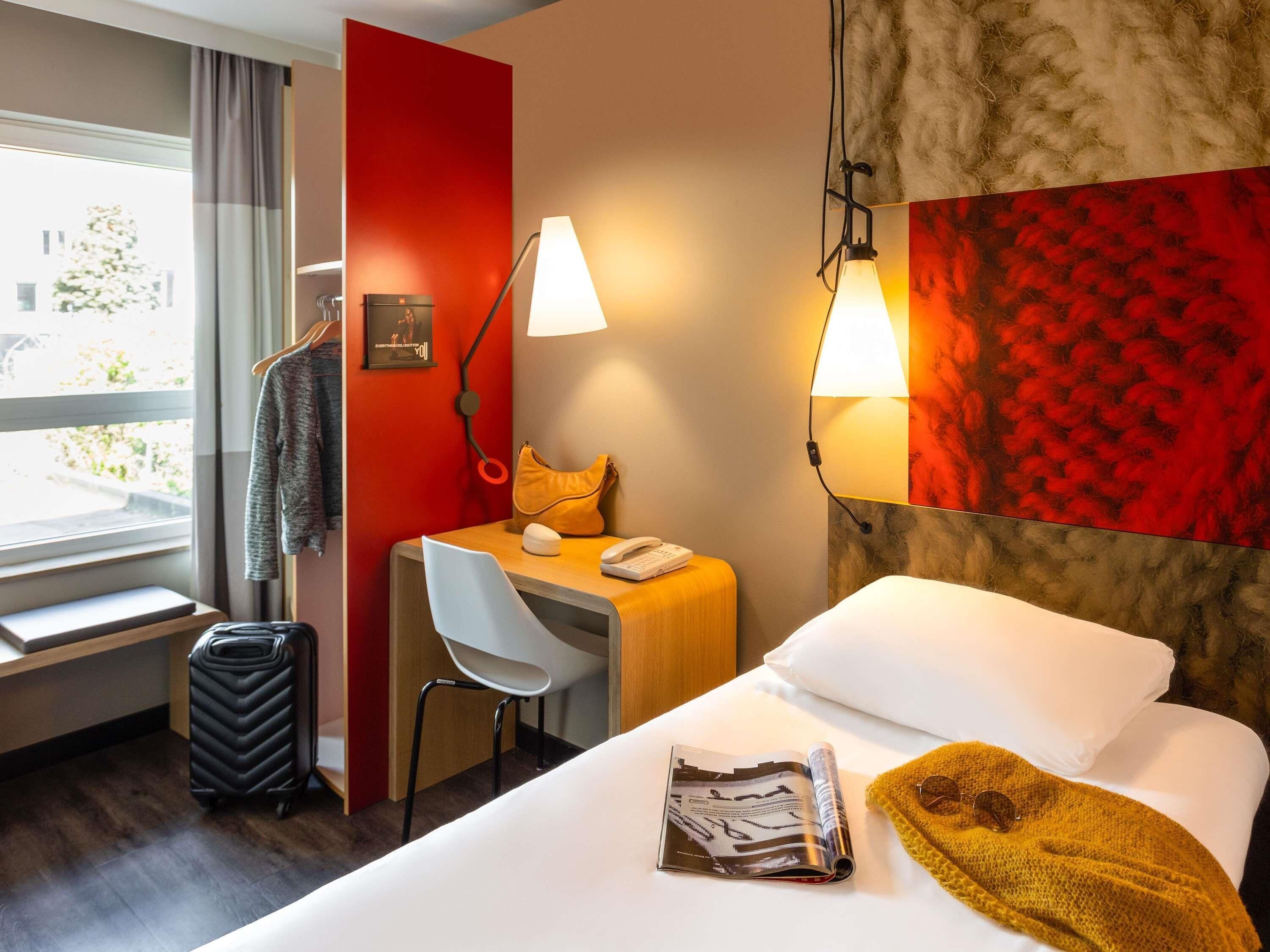 Habitación de hotel con cama, un escritorio, una silla, una maleta, una lámpara y una ventana con cortinas.
