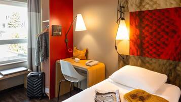 Habitación de hotel con cama, un escritorio, una silla, una maleta, una lámpara y una ventana con cortinas.