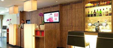 A hotel reception area with a wooden reception desk, a television screen, and a bar with various bottles.