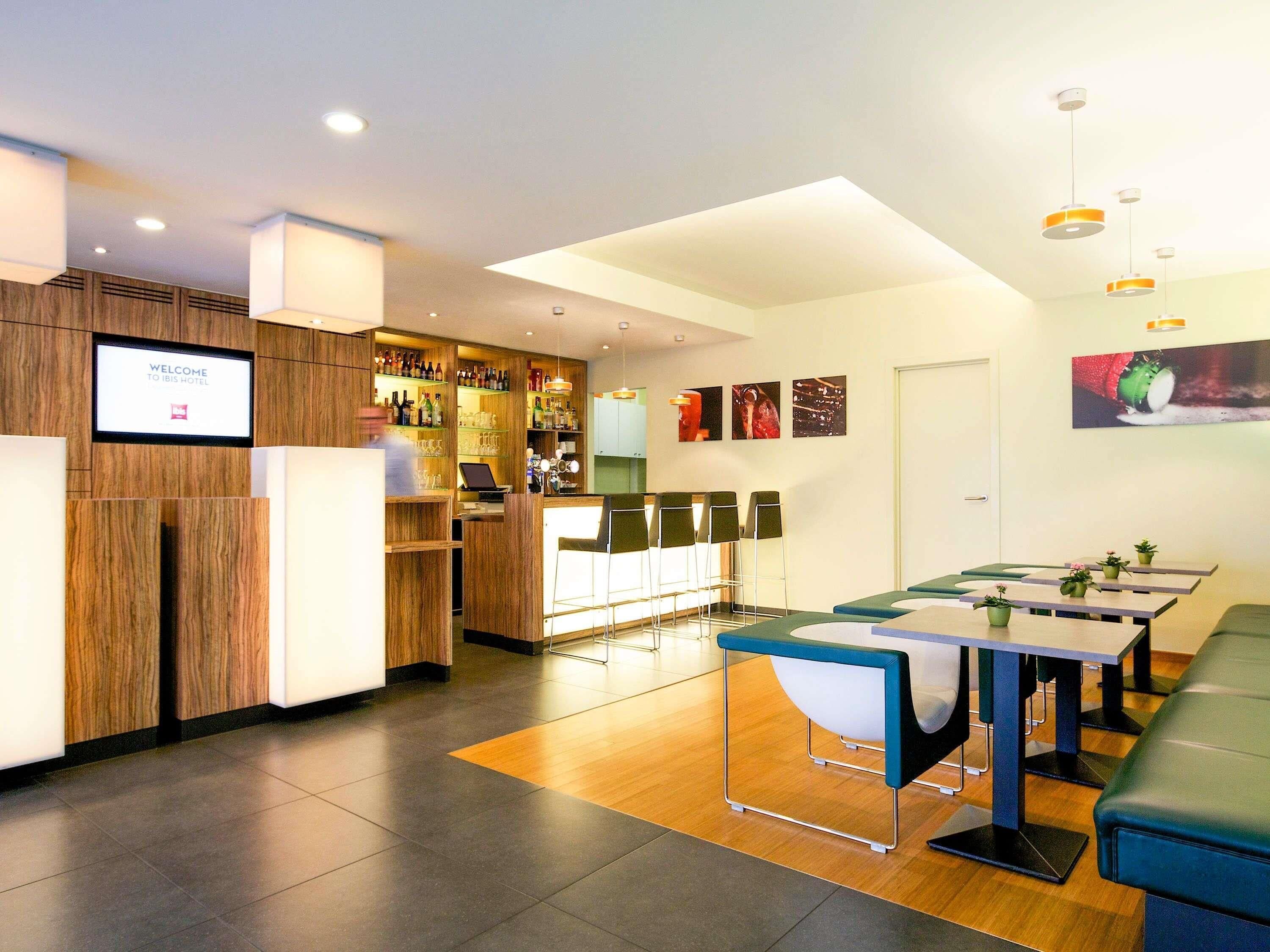 A modern hotel lobby with a reception desk, bar area, seating, and a digital welcome screen.