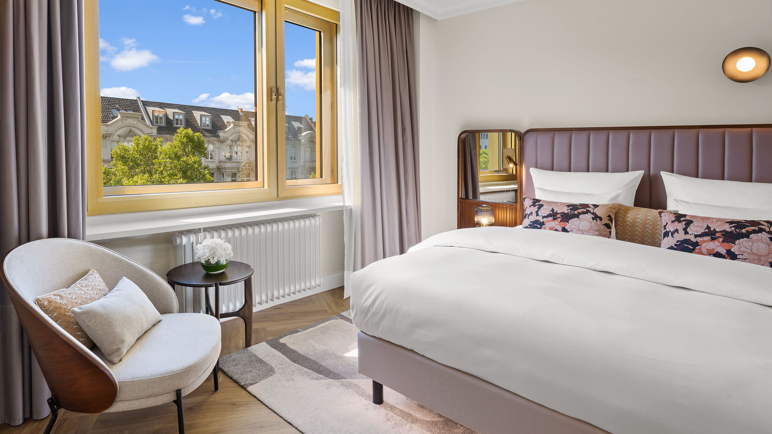 A hotel room with a large bed, a chair, a small table with a vase, a window with curtains, and a view of buildings outside.