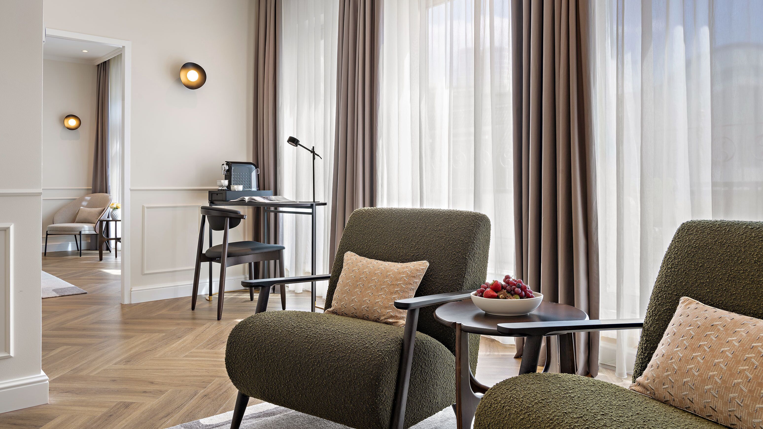 Ein modernes Wohnzimmer mit zwei grünen Sesseln, einem kleinen Tisch mit Obstschale und einem Schreibtisch mit Laptop und Lampe.