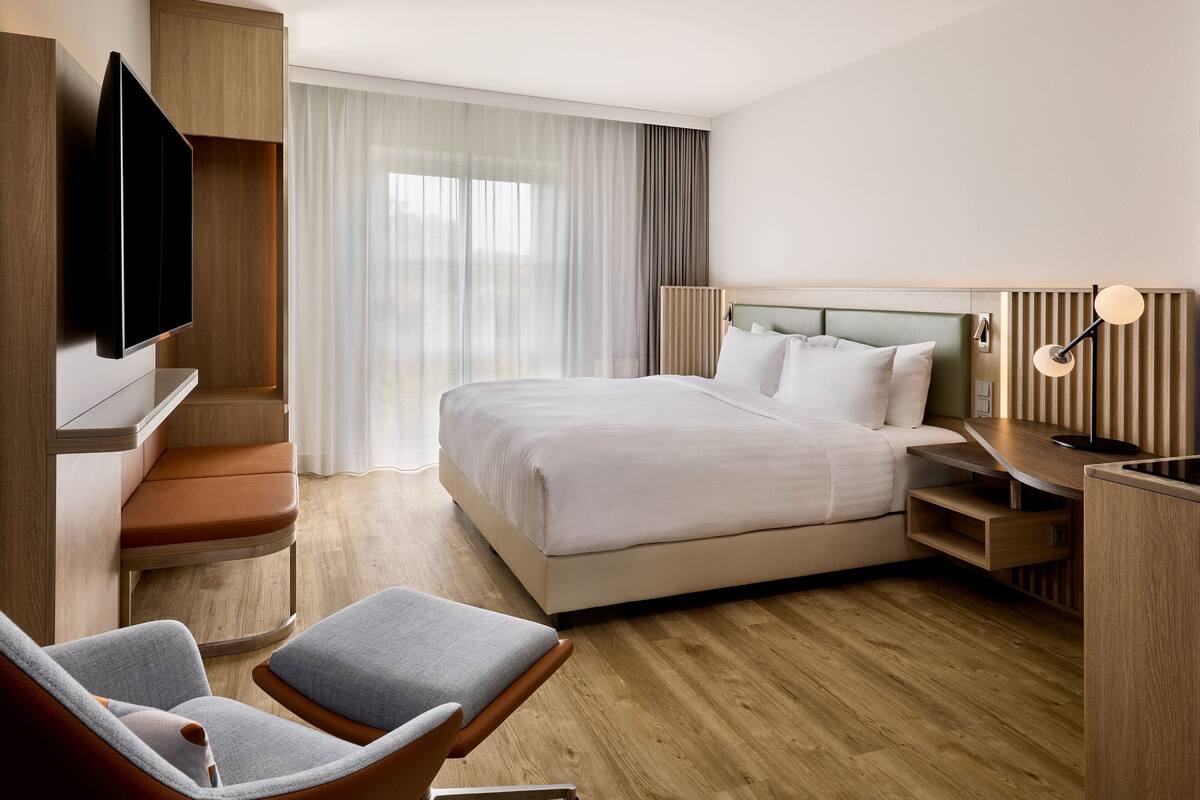 Une chambre d’hôtel moderne dotée d’un grand lit, d’un téléviseur à écran plat fixé au mur, d’un coin salon avec un fauteuil et un pouf, et d’un bureau en bois.