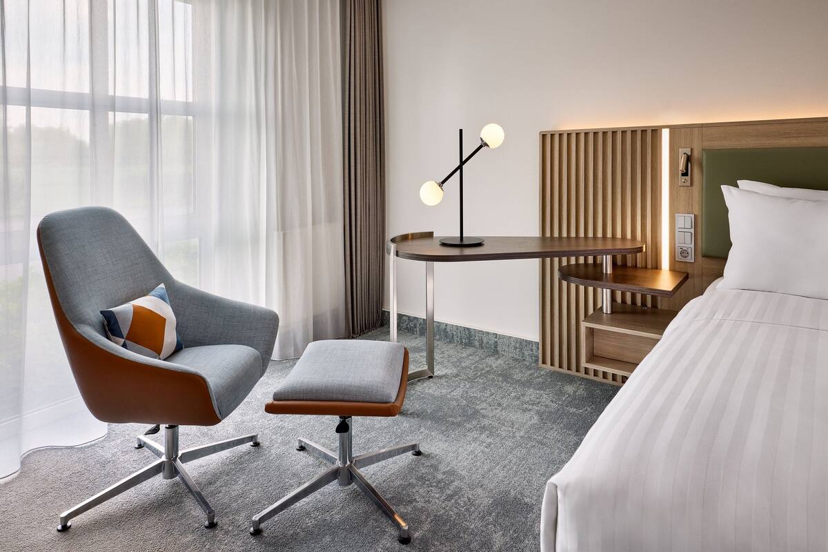 Une chambre d’hôtel moderne avec un fauteuil à revêtement gris, une petite table d’appoint en bois, une lampe de bureau et un lit avec du linge de lit blanc.