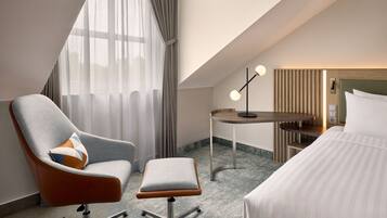 A hotel room with a modern design, featuring a large bed, a seating area with a chair and ottoman, a small table, and a large window with curtains.
