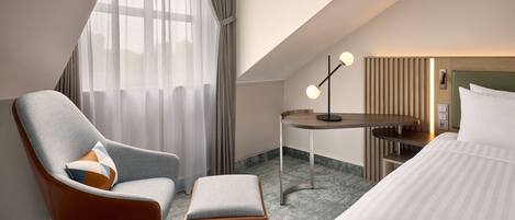 A hotel room with a modern design, featuring a large bed, a seating area with a chair and ottoman, a small table, and a large window with curtains.