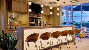A bar area with a long counter, multiple stools, and a variety of bottles. - Courtyard by Marriott Magdeburg (Barleben)