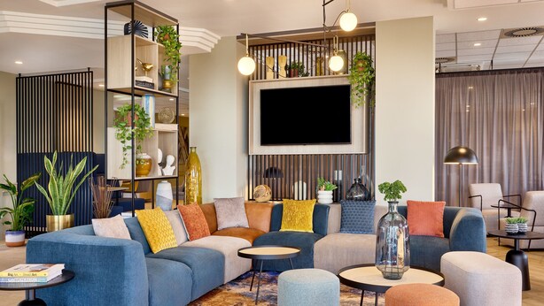 A modern living room with a sectional sofa, ottomans, a coffee table, and a TV wall-mounted above a wooden panel. - Courtyard by Marriott Magdeburg (Barleben)