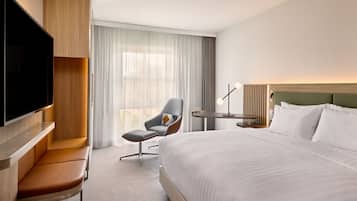 Una habitación de hotel moderna con una cama grande, un televisor de pantalla plana, una zona de estar con una silla y reposapiés, una mesita pequeña y un ventanal con cortinas.