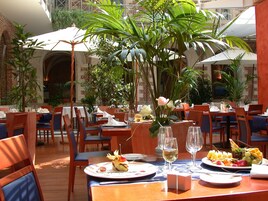 Un área de comedor al aire libre con mesas dispuestas para comidas, rodeada de plantas en macetas y sombrillas.